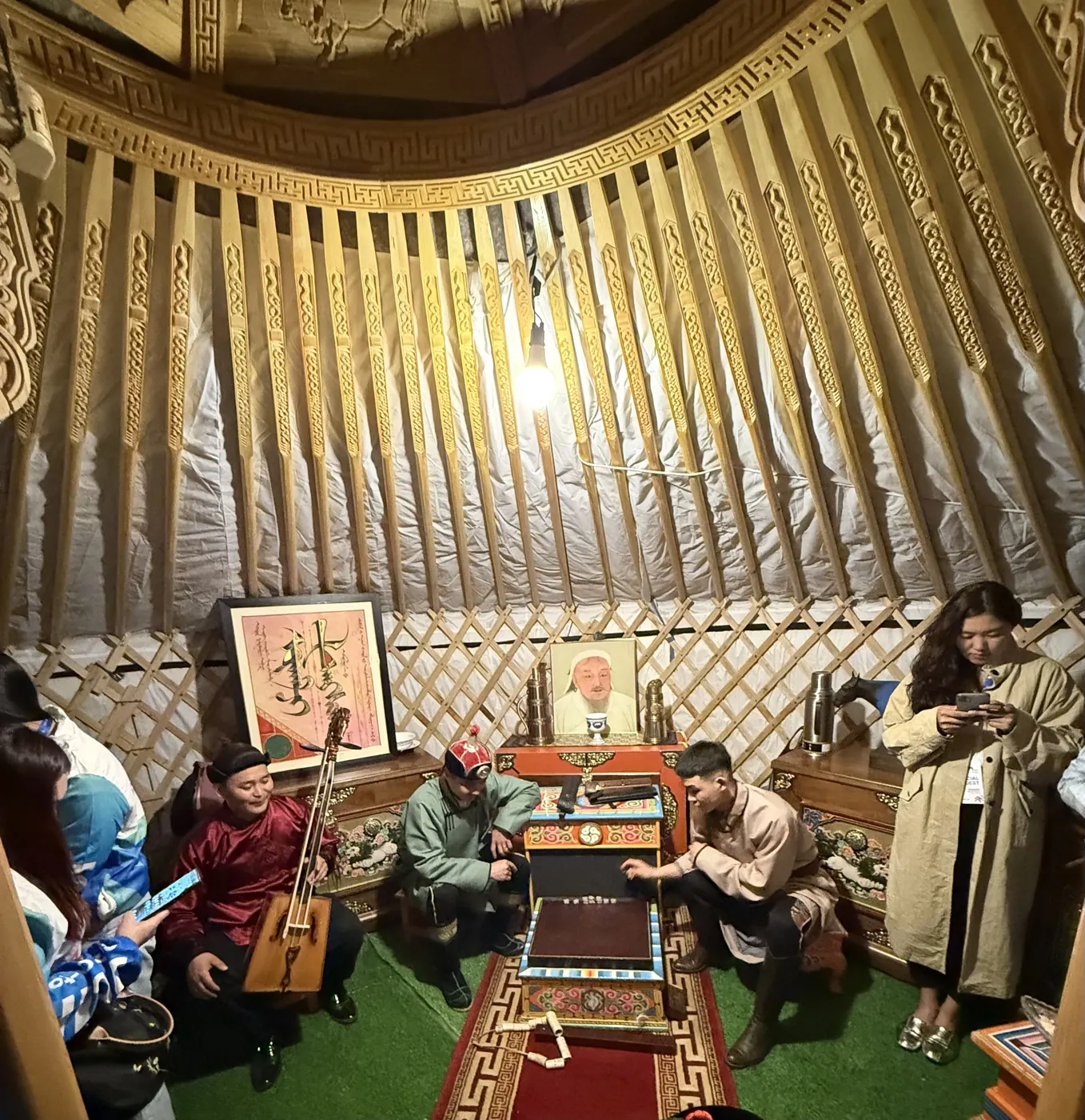 Musicians in deel robes, one holding a horsehead fiddle, inside a ger with a portrait of Genghis Khan