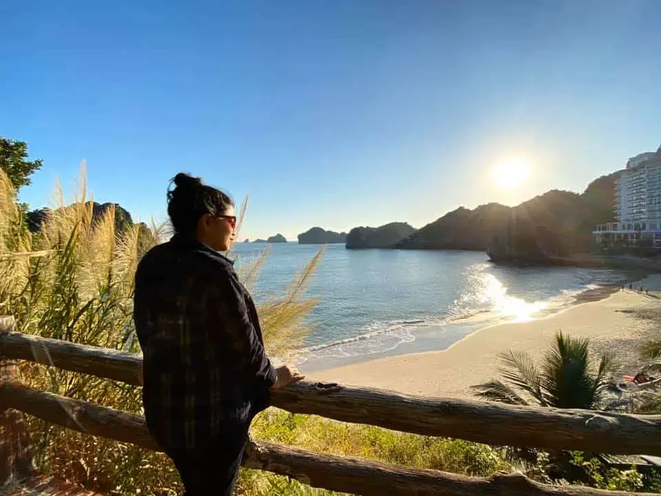 Afshana leaning on a wooden rail above a beach on Cat Ba Island in the morning sun